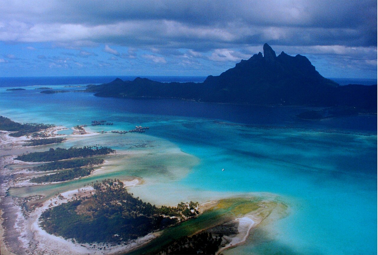 Vue aérienne du lagon de Bora Bora, avec des bungalows sur pilotis au premier plan et le mont Otemanu au fond