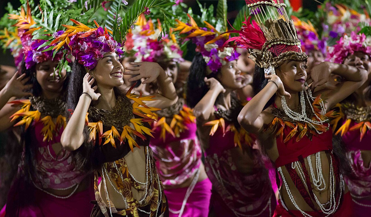 Danseuses souriantes en couronnes de fleurs violettes et colliers de coquillages, en tenue rose, lors d'un spectacle de nuit