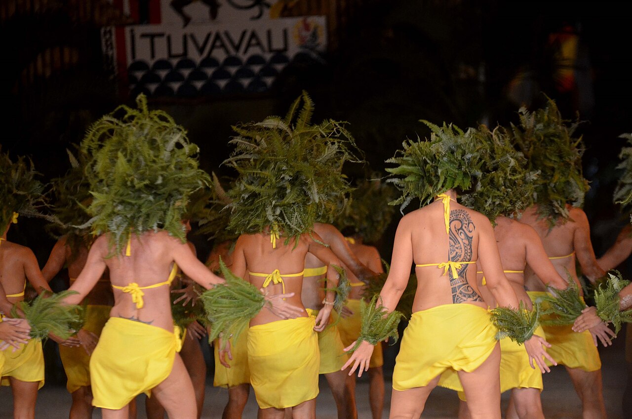 Danseurs de dos en paréos jaunes et coiffes de fougères vertes sur scène, lors d'une soirée de Heiva