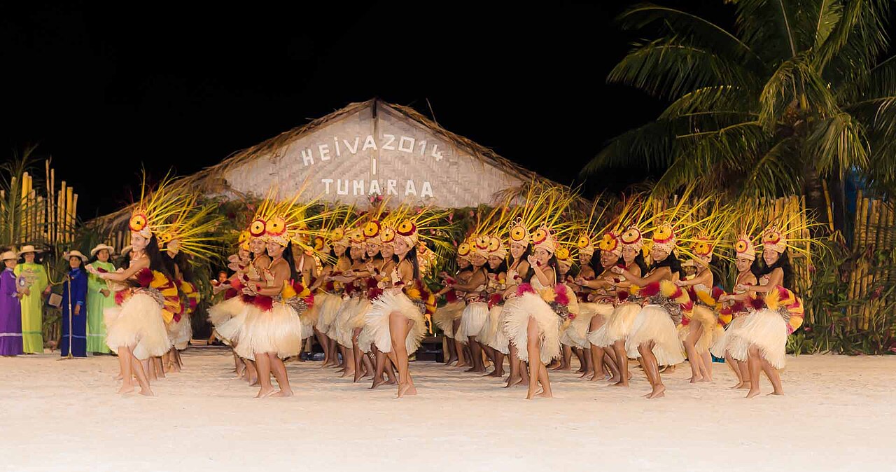 Danseuses en costumes de fibres et coiffes jaunes et rouges sur une scène de sable, de nuit, lors du Heiva 2014