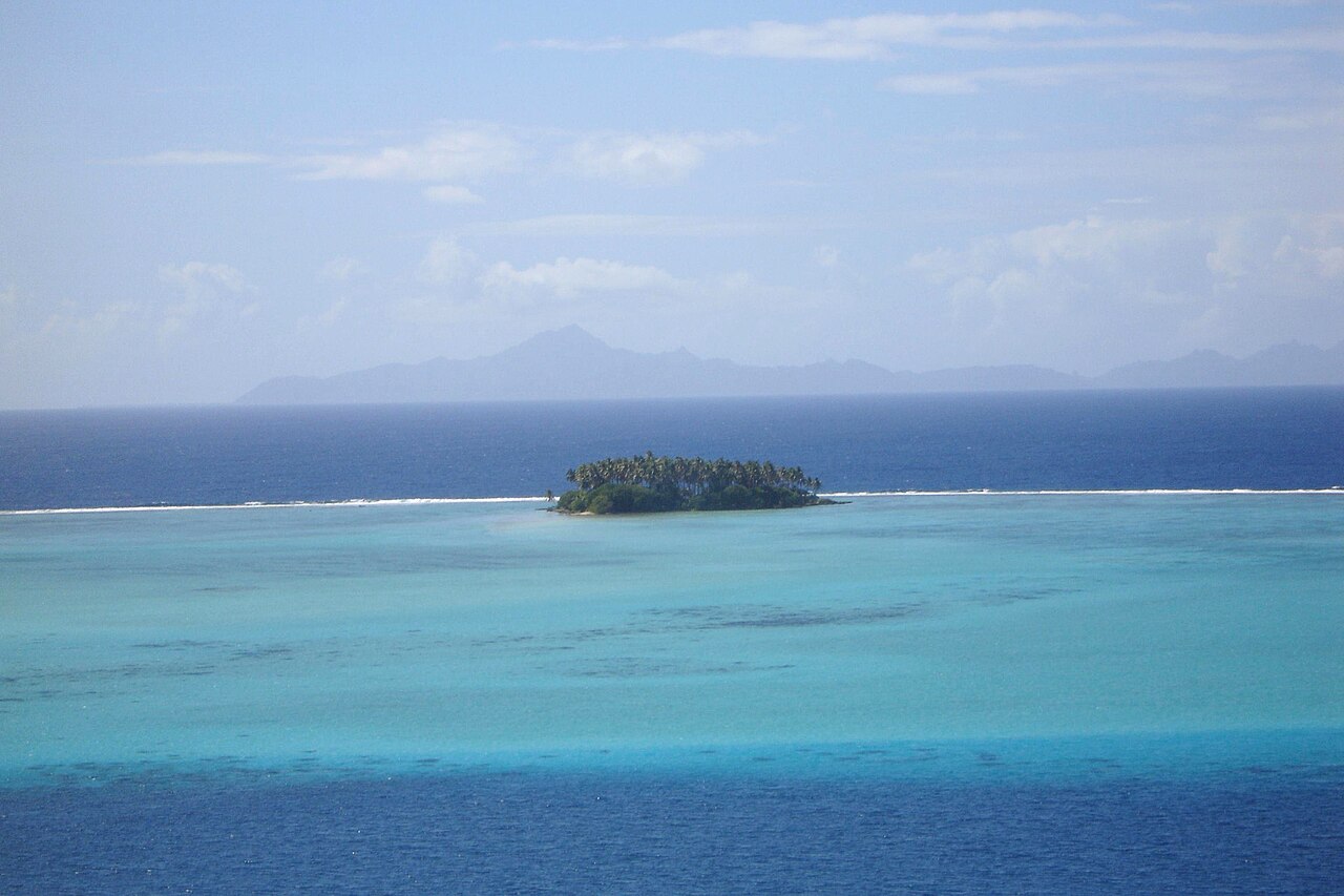 Petit motu couvert de cocotiers au milieu d'un lagon turquoise, avec une île montagneuse à l'horizon