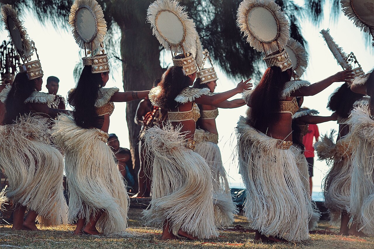 Danseuses tahitiennes en tenue traditionnelle, de dos, avec coiffes ornées de nacre et jupes de fibres, sur une pelouse au bord de la mer