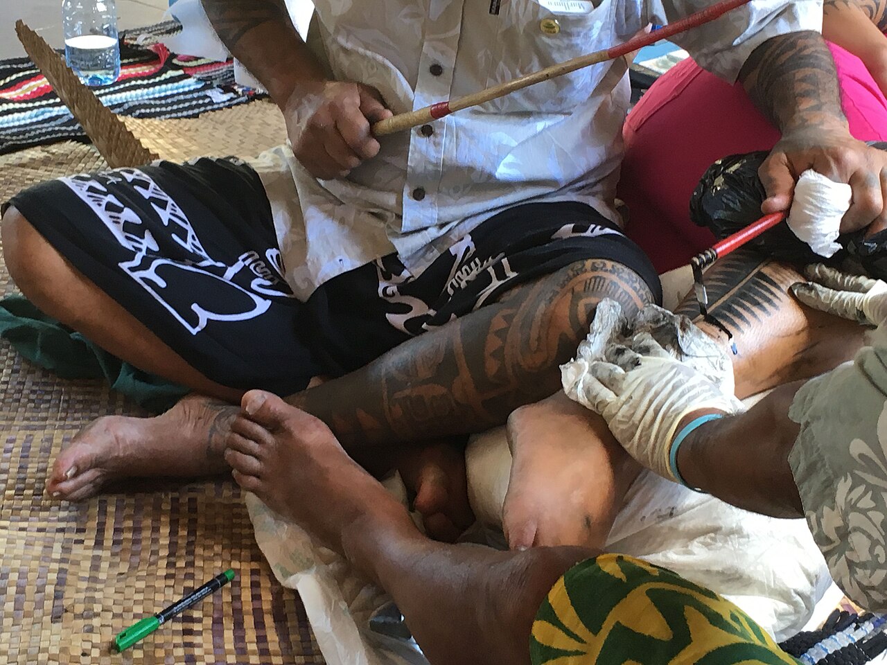 Tatouage traditionnel polynésien réalisé à la main avec un peigne et un bâtonnet, sur la jambe d'une personne assise sur une natte
