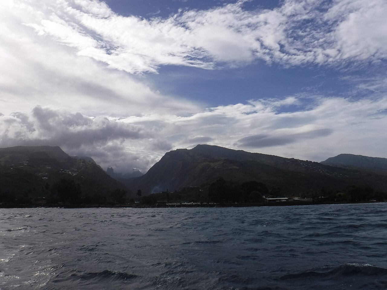 Vue depuis la mer sur la baie de Punaauia : montagnes verdoyantes de Tahiti, côte bordée d'arbres et ciel nuageux