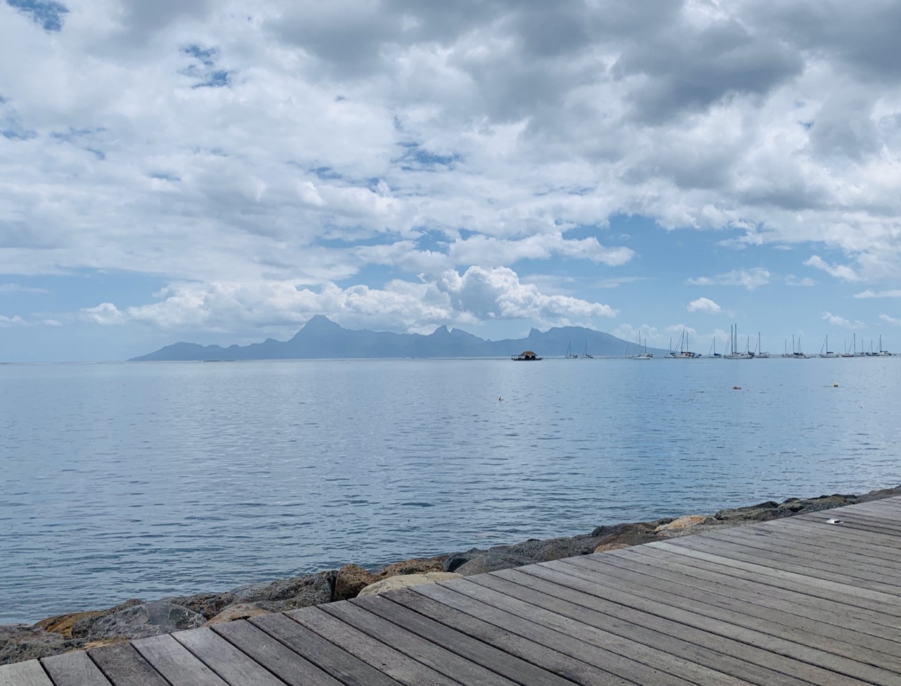 Front de mer de Punaauia avec un ponton en bois, la mer calme et l'île de Moorea à l'horizon sous un ciel nuageux