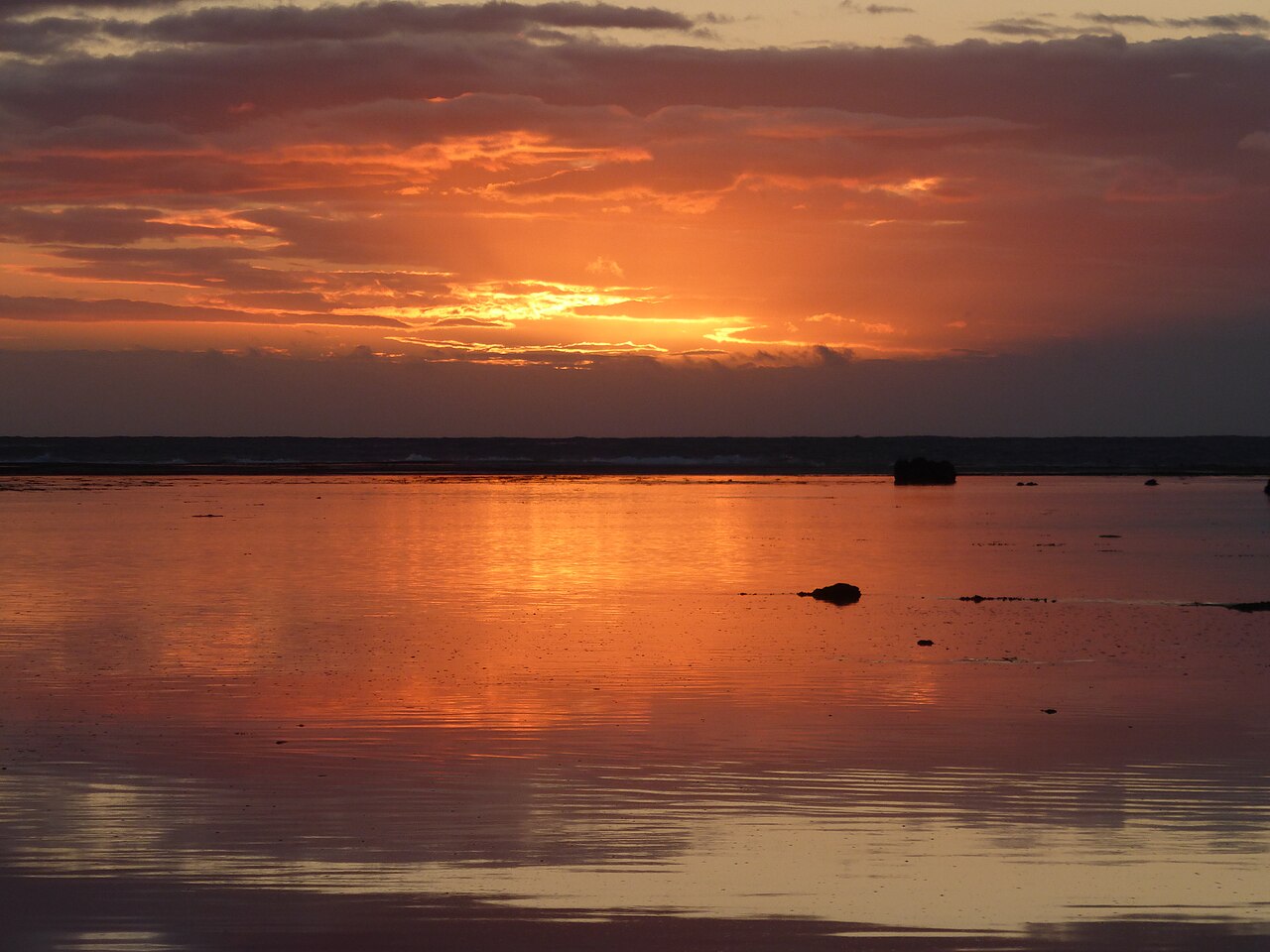 Coucher de soleil orangé qui se reflète sur le lagon calme d'une plage de Punaauia, à Tahiti